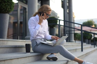 Sırıtkan sarışın kadın dışarıda otururken dizüstü bilgisayar kullanıyor ve akıllı telefonla konuşuyor.