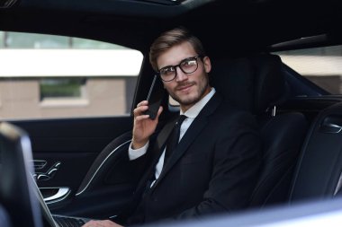 Yakışıklı işadamı arabanın arka koltuğunda dizüstü bilgisayar ile oturan telefon ile konuşuyor