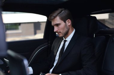 Takım elbiseli yakışıklı, akıllı telefonla konuşan ve arabada otururken dizüstü bilgisayar kullanarak çalışan yakışıklı kendine güvenen bir işadamı