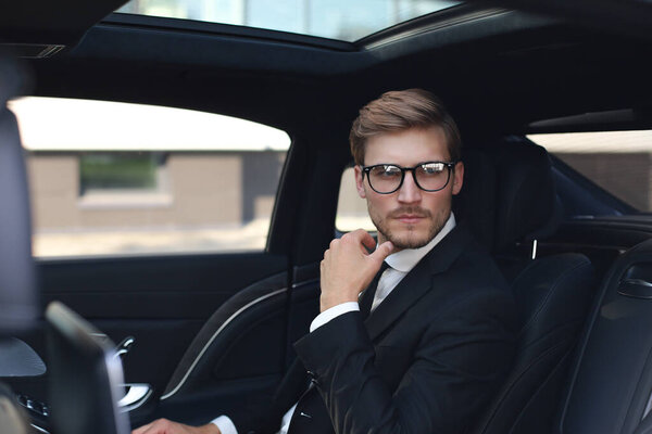 Thoughtful confident businessman in full suit with eyewear looking away while sitting in the car