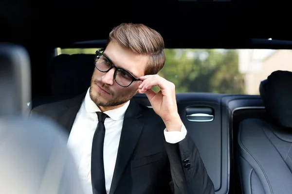 Thoughtful confident businessman keeping hand on glasses while sitting in the luxe car