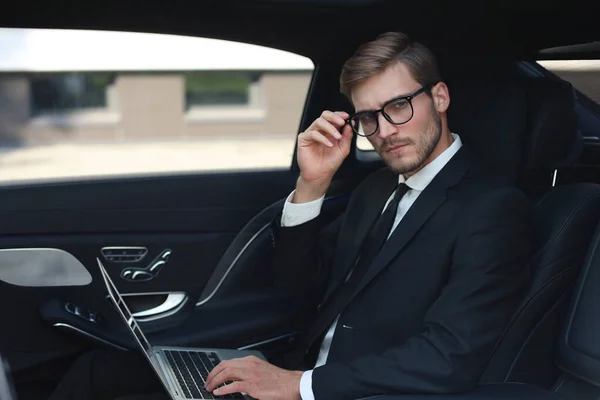 Thoughtful confident businessman keeping hand on glasses while sitting in the luxe car and using his laptop