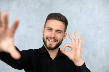 Gri arkaplanda izole edilmiş selfie çekilirken tamam işareti gösteren neşeli iş adamı görüntüsü.