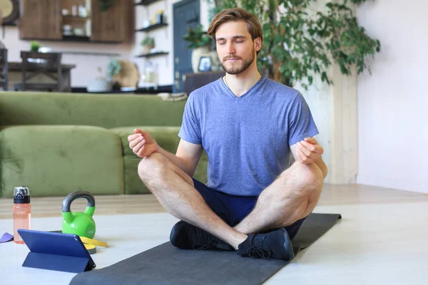 若いハンサムな男は隔離中に自宅で床に座ってハスのポーズでヨガを練習しています スポーツは健康的な生活の基盤です — ストック写真