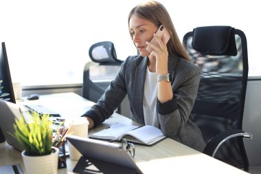 Güzel bir iş kadını modern ofiste otururken cep telefonuyla konuşuyor.