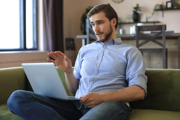 stock image Concentrated young freelancer businessman sitting on sofa with laptop, working remotely online at home