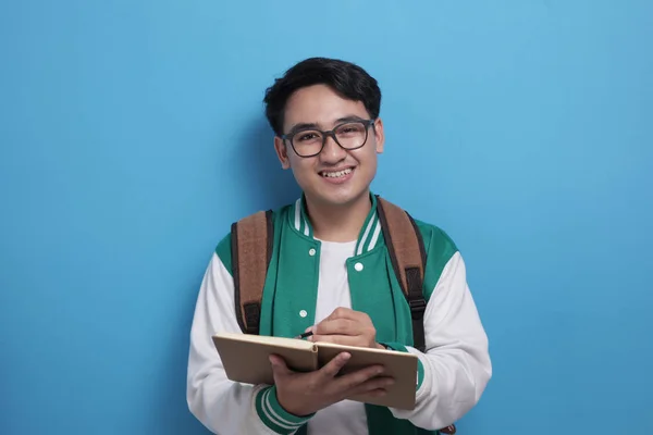 Joven estudiante asiático vistiendo chaqueta de béisbol verde escribiendo en libro — Foto de Stock