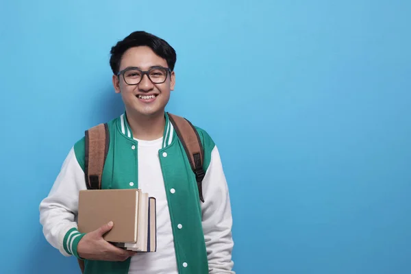 Junger asiatischer Student in grüner Baseballjacke, Bücher in der Hand und lächelnd in die Kamera — Stockfoto
