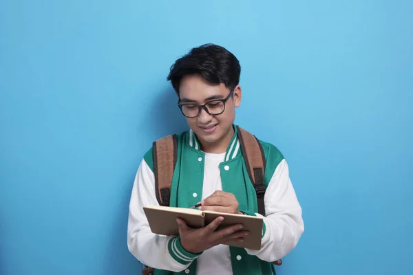 Giovane asiatico maschio studente indossare verde giacca da baseball scrittura su libro — Foto Stock
