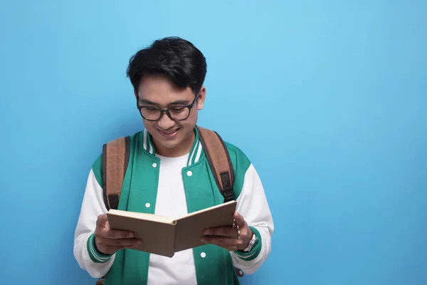 Maschio asiatico studente sorridente e la lettura di un libro contro blu sfondo — Foto Stock