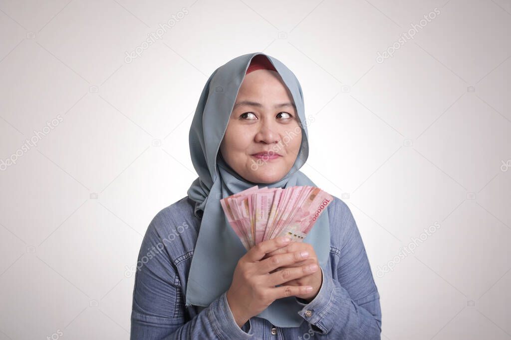 Portrait of Indonesian muslim woman holding rupiah money, smiling and thinking gesture