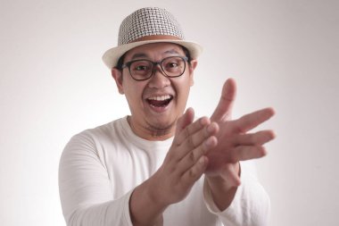 Beyaz tişörtlü genç Asyalı adam, mutlu bir alkış hareketi. Gri arka planda, yarım vücut portresi üzerinde.