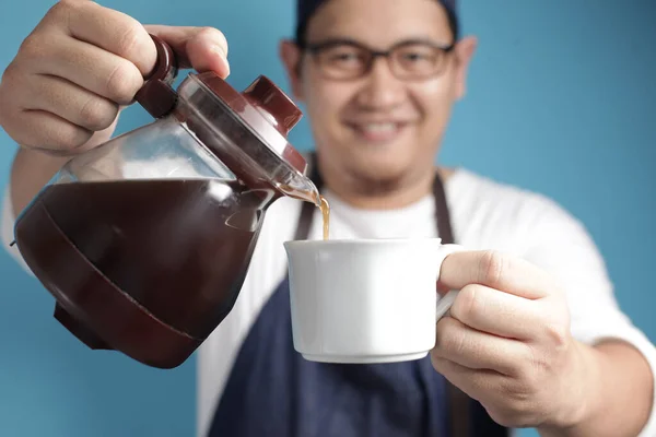 Portrait of Asian male chef or waiter smiling while pouring coffee to a cup, offering coffee concept, against blue background