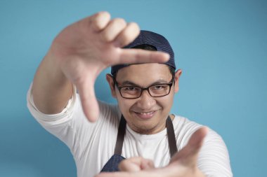 Asyalı erkek aşçının ya da garsonun portresi kameraya bakıyor ve yüzünü mavi arka planla çerçevelerken gülümsüyor