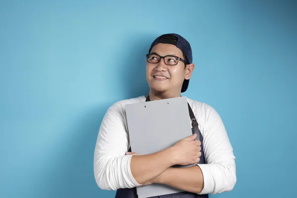 Portrait of Asian male chef or waiter smiling and thinking while holding clipboard, successful entrepreneur in good restaurant business concept, against blue background