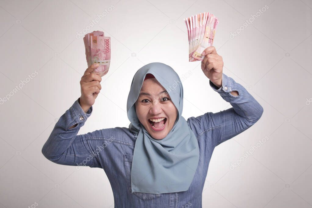 Portrait of Indonesian muslim woman holding rupiah money, smiling laughing winning gesture