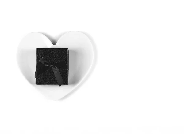 Caja de regalo negra con lazo, un plato blanco en forma de corazón sobre fondo blanco. Concepto minimalista negro — Foto de Stock