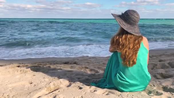 Mulher bonita usando um chapéu sentado em uma praia de areia e olhando para o oceano azul. Menina em vestido turquesa olhando para a distância, humor de verão. Relaxa na praia. Florida, EUA — Vídeo de Stock