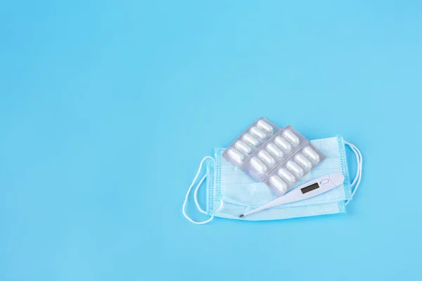 Face mask, thermometer, pills on a blue background. Medicine supplies for home coronavirus COVID-19 quarantine. Space for copy. — Stock Photo, Image