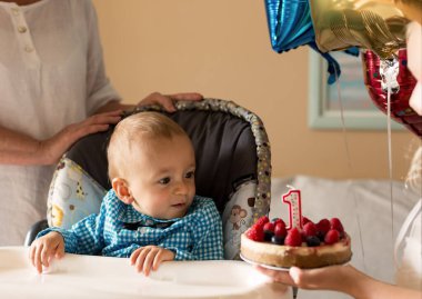 Bir yaşında tatlı bir çocuk ve mumlu dutlu kek. Kutlama tişörtü giyen, sandalyeye oturan bir çocuk. Evde ilk doğum günün kutlu olsun. Aile kavramı.