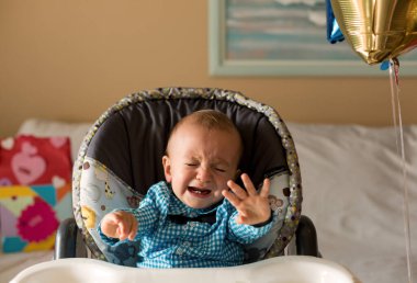 Bir yaşında tatlı bir çocuk ağlıyor. Birinci doğum günün kutlu olsun. Kutlama tişörtü giyen bir çocuk, sandalyede oturuyor. 