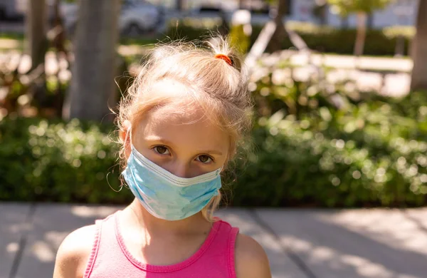 Küçük bir kız tıbbi maskeyle parkta yürüyor. Küçük bir kız boş bir şehirde yürüyor ve yüzünü bir koronavirüs maskesiyle örtüyor. Coronavirus karantinası zamanı. Boşluğu Kopyala. Stok Fotoğraf