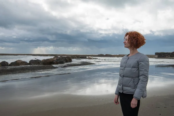 happy young woman in puffer jacket on seashore on cloudy day, Anzio, Italy