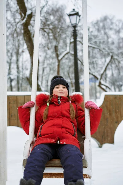 Zimní portrét malé holčičky v klobouku a červené bundě na houpačce — Stock fotografie