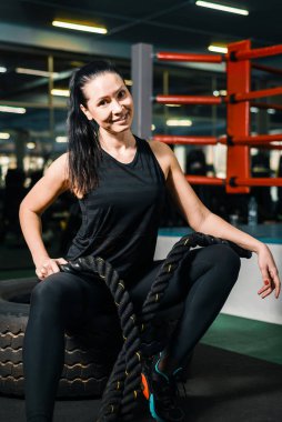 Spor salonundaki güzel bir kızın portresi. Güzel bayan gülümsüyor. Benekli kız ipleri elinde tutuyor. Ellerinde antrenman yapıyor ve lastiğe oturuyor.