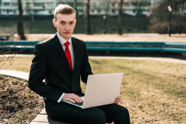 Parktaki bankta dizüstü bilgisayarıyla oturan genç bir iş adamı. Kırmızı kravatlı takım elbiseli adam. Serbest çalışıyorum. uzaktan çalışma konsepti. Y nesli, milenyum