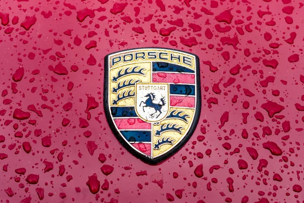 Motorkap embleem van sportieve auto Porsche in regendruppels op de Bourgondische achtergrond. — Stockfoto