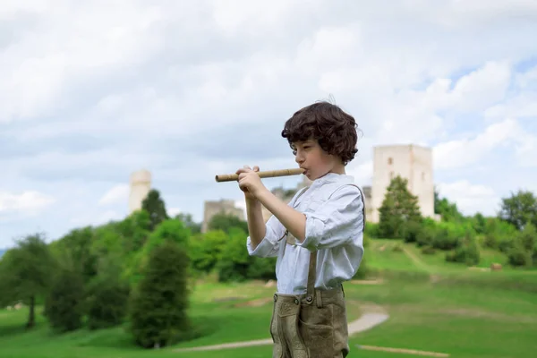 A boy in traditional Bavarian clothes with a shepherd's pipe on the background of ancient ruins. — Stock Photo, Image