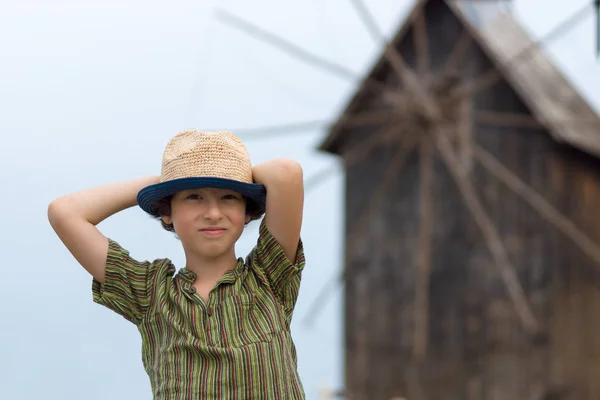 Portrait of a beautiful boy in a short shirt and hat on a blurred background of a wooden windmill. — Stock Photo, Image