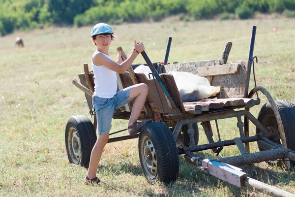 A little boy in a T-shirt, shorts and a cap near an old cart.