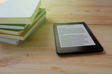 E-kitap okuyucu ve ahşap masada yığınla kitap.