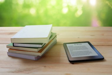 E-kitap okuyucu ve ahşap masada yığınla kitap.