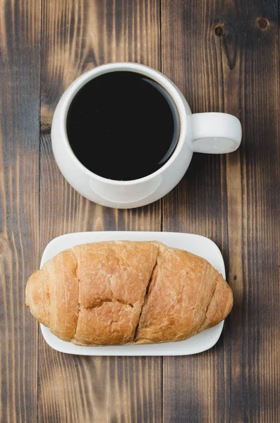 Caneca branca com café preto e croissant no fundo de madeira . — Fotografia de Stock