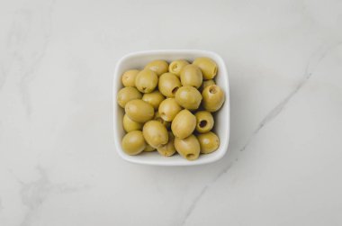 Beyaz taşlı arka planda bir kasede yeşil zeytin turşusu. Üst 