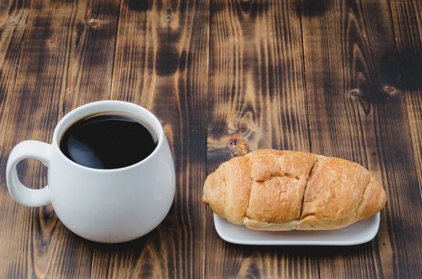 Caneca branca com café preto e croissant no fundo de madeira . — Fotografia de Stock