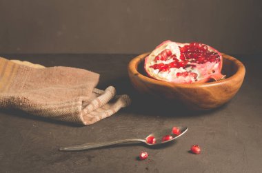 Ahşap bir kâse içinde kırmızı bir kepçeyle kesilmiş tahta bir kâse, tahtadan bir kâse içinde nar parçaları ve koyu bir arka planda bir kaşık, seçici bir odak noktası.