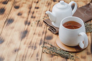 Beyaz fincanda kekik çayı ve ahşap masada çaydanlık, metin için yer.