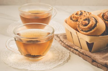 Tahta bir sepetin içinde haşhaş ile iki bardak çay ve tahta sepetin içinde haşhaş ile beyaz bir masada seçici bir odak noktası olan tahta bir sepetin içinde rulo.