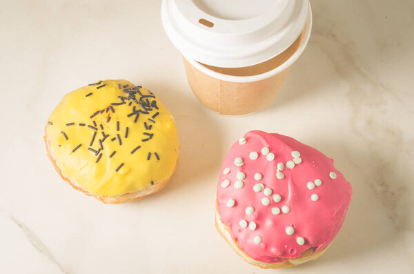 два пончика в глазури и бумажная чашка кофе / два пончика в глазури и бумажная чашка кофе на белом мраморном фоне. Вид сверху
