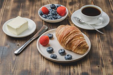 Kahve fincanı, kruvasan, beyaz kasede böğürtlen ve ahşap arka planda tereyağı. Taze böğürtlenli sağlıklı kahvaltı