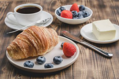 Kahve fincanı, kruvasan, beyaz kasede böğürtlen ve ahşap arka planda tereyağı. Taze böğürtlenli sağlıklı kahvaltı