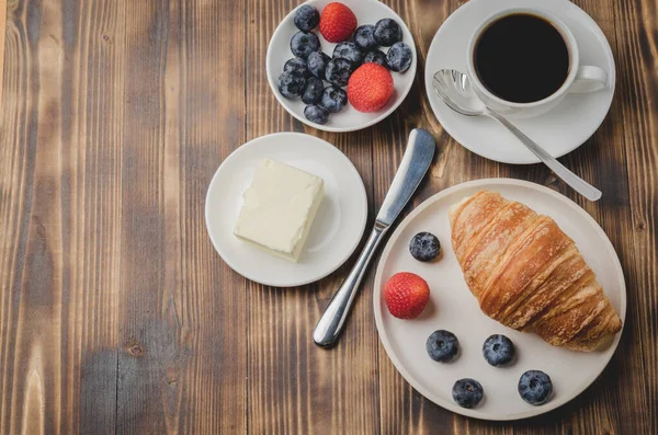 Coffee cup, croissant with berries in white bowl and butter knife on wooden table. Top view and copyspace. Healthy breakfast with fresh berries