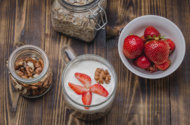 Sağlıklı kahvaltı. Yoğurt, taze çilek, ev yapımı granola ve ahşap bir masada açık cam kavanozda ceviz. Üst yaşam