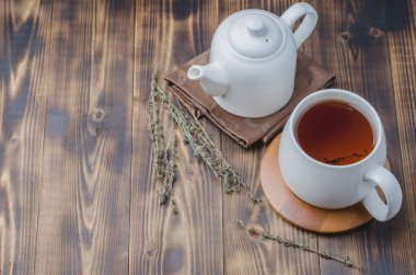 Beyaz fincanda kekik çayı ve ahşap masada çaydanlık, metin için yer.