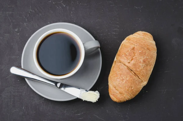 Kruvasanla kahve molası, tereyağı bıçağı ve koyu bir masada bir fincan sade kahve. Üst görünüm.