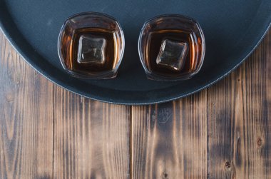 Siyah tepside buzlu viski bardakları ve ahşap masa / Siyah tepside buz küpleri olan iki bardak viski ve kopyalanmış ahşap masa. Üst görünüm.